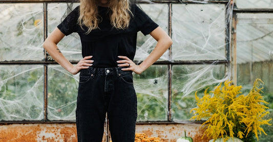 A woman dressed as a witch stands among pumpkins in a greenhouse, surrounded by spider webs.