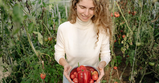 Woman smiling while holding a bowl of fresh tomatoes and peppers in a lush greenhouse.