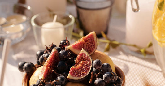 A wooden bowl filled with grapes and figs, surrounded by lit candles on an outdoor table setting.