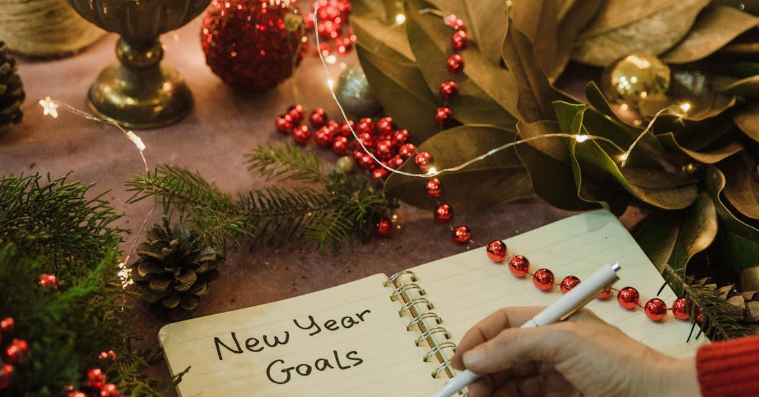 Hand writing New Year goals in a festive notebook surrounded by Christmas decorations and fairy lights indoors.