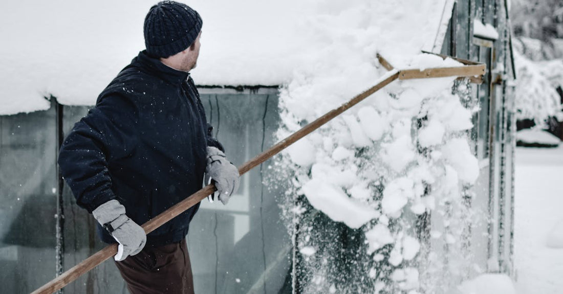 Man using a roof rake to remove snow from a greenhouse roof during a cold winter day in Estonia.