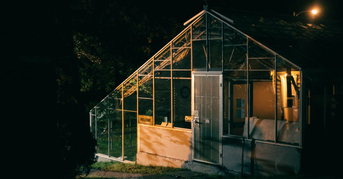 A warmly lit greenhouse exterior with glass panels illuminated at night.